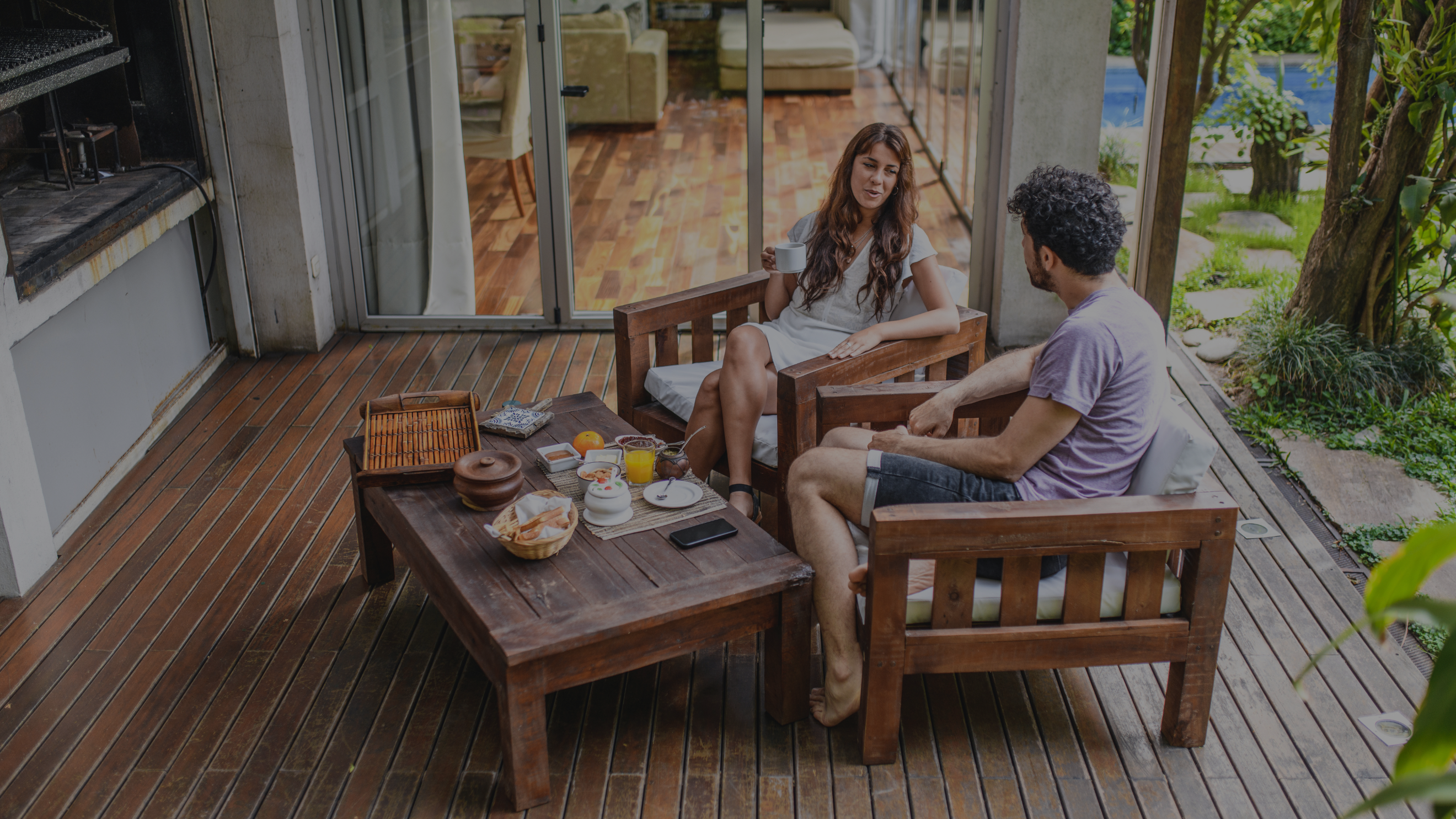 Two people sitting on a wooden deck with a table and chairs, surrounded by greenery.